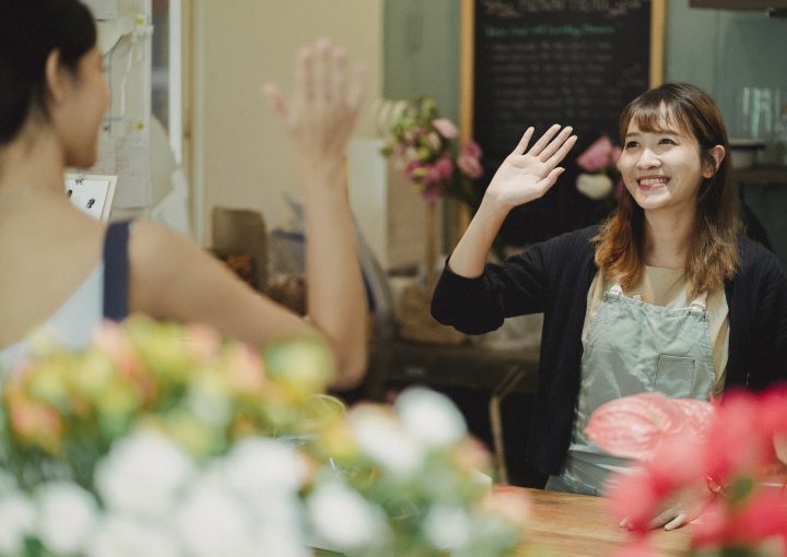 Positive Asian woman greeting client waving hand while working as florist in flower shop